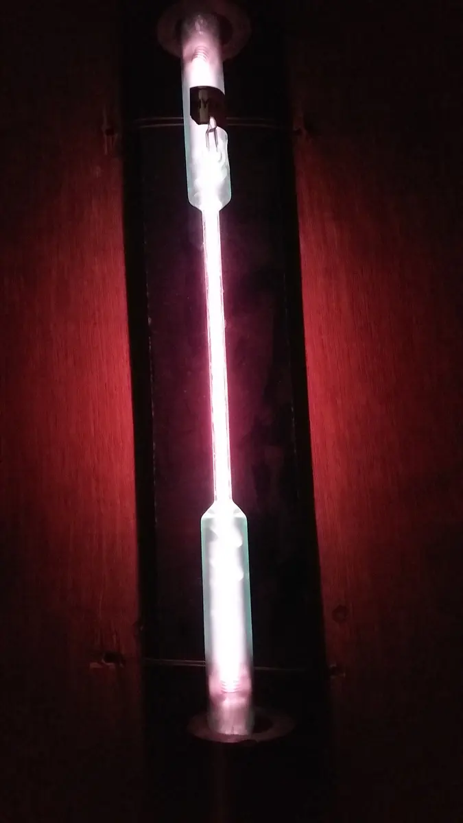 A thin upright glass tube glowing bright pinkish white in a dark room, with metal electrodes at its ends