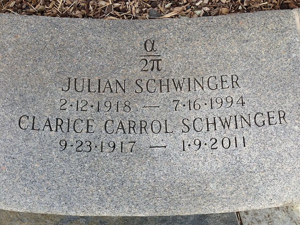A flat gray granite gravestone on the ground, carved with the symbols alpha over 2 pi, and below them the names and dates of Julian Schwinger, 1918 to 1994, and Clarice Carrol Schwinger