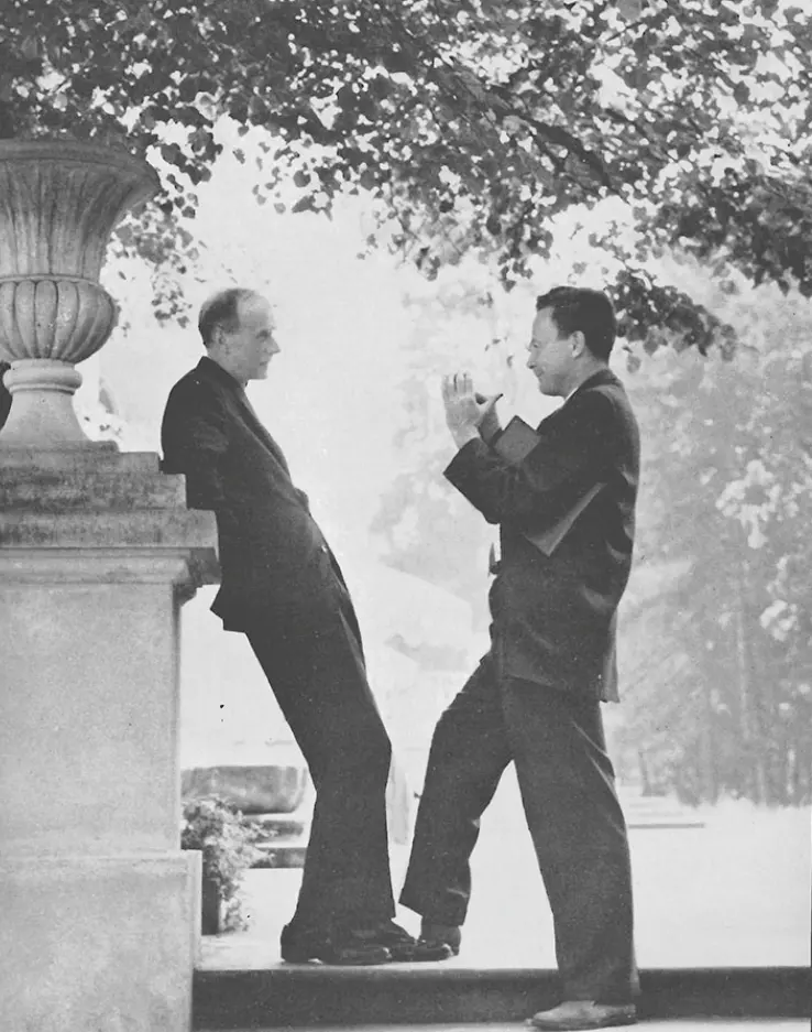 Black-and-white photo of two men in dark suits outdoors beside a stone wall under trees: one leans back against the wall listening, the other stands facing him, talking with both hands raised