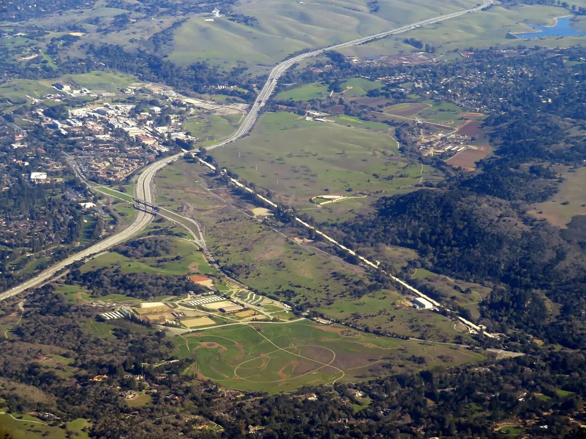 An aerial photo of green hills with a long, perfectly straight line running across them towards a cluster of buildings, with a motorway crossing it
