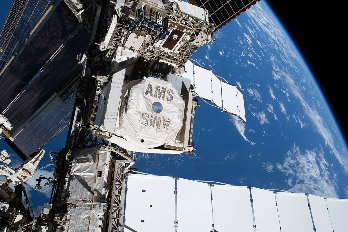 A large boxy instrument marked AMS fixed to the metal frame of the International Space Station, with the blue Earth and white clouds below
