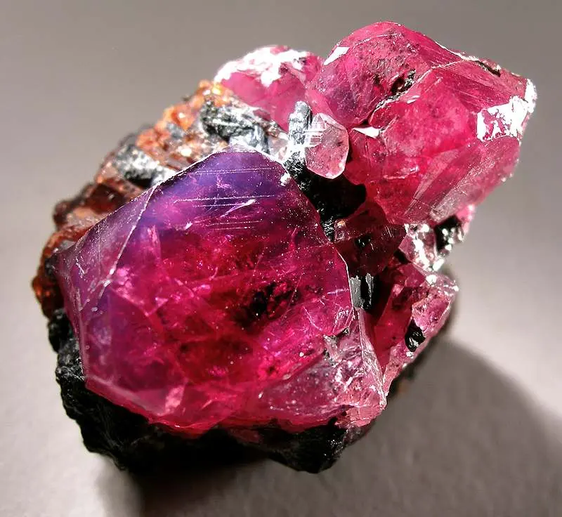 A rough, glassy crystal, deep pinkish red, with dark rock at its base
