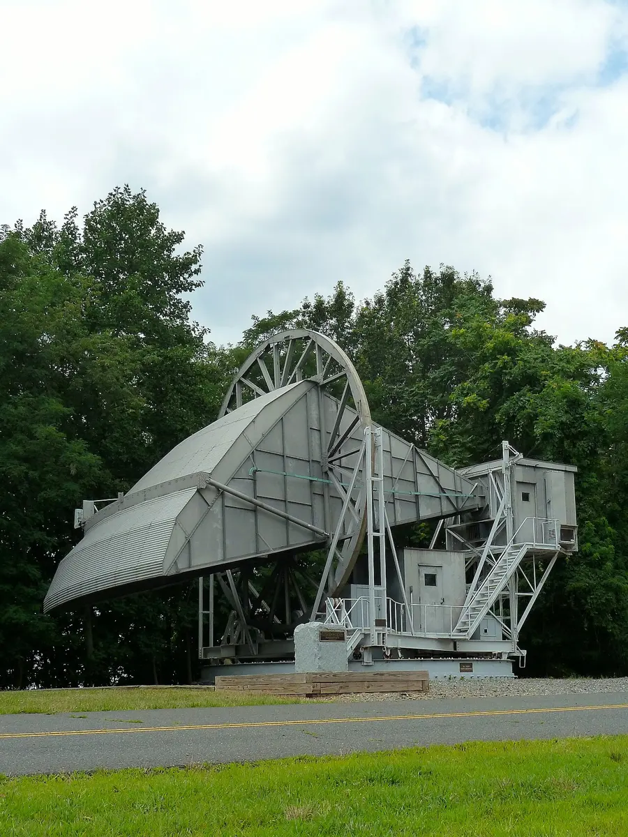 A huge gray metal antenna shaped like a funnel lying on its side, on a tall metal frame with stairs, among green trees beside a road