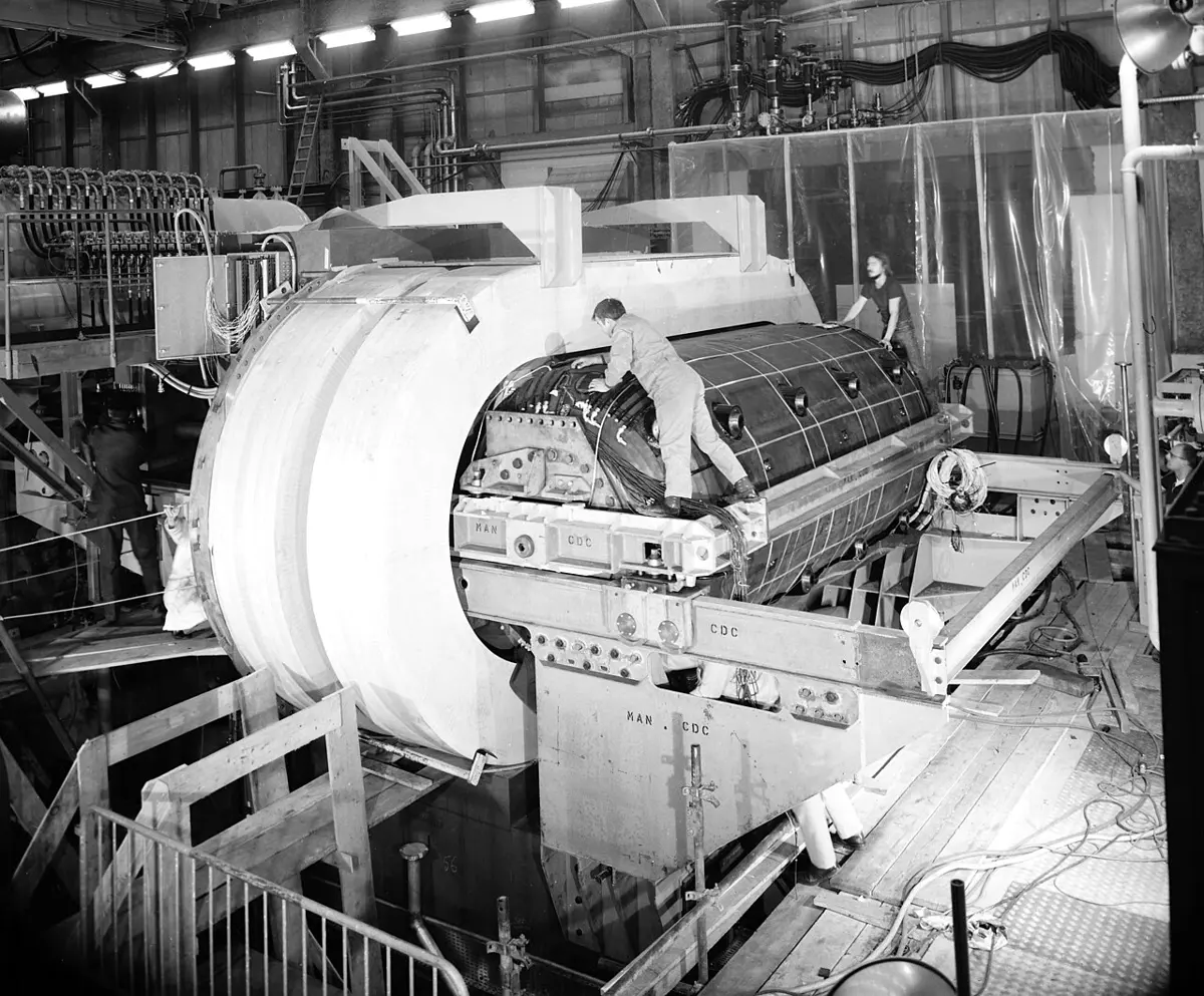 A black and white photo of a huge machine in a large hall: a long cylinder, with workers climbing on it, being slid into a big white ring-shaped magnet