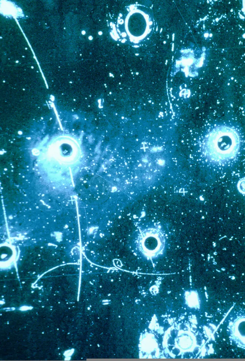 A dark blue photo full of bright round spots, the chamber’s lamps and windows, crossed by a few thin, pale curving lines, the particle tracks