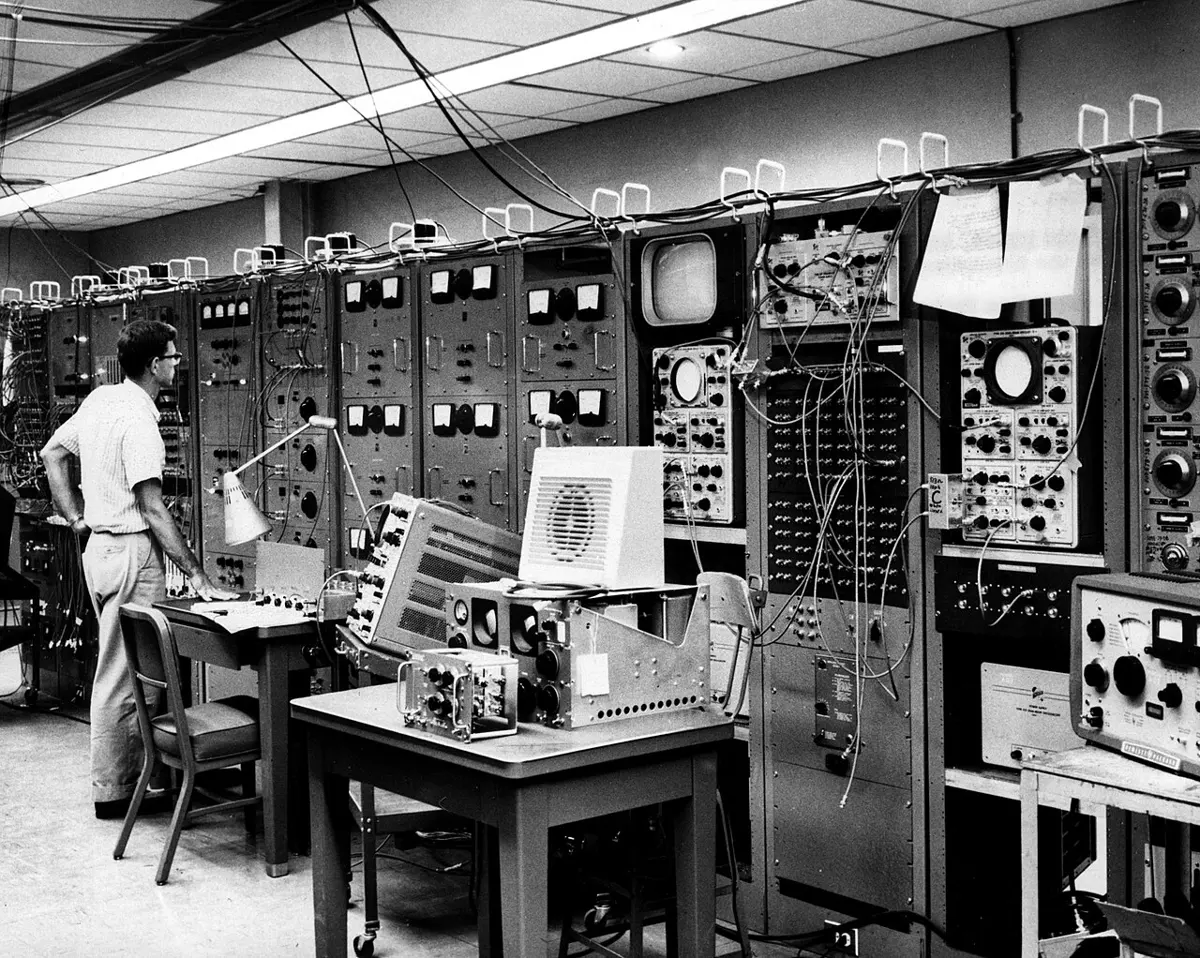 A man in a short-sleeved shirt standing at a desk in a long room lined with tall racks of electronic equipment, dials, switches, screens and cables