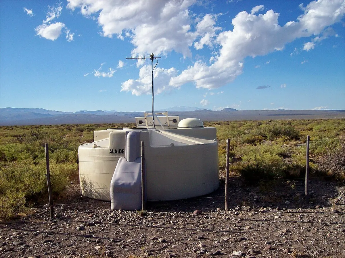 A large round cream-colored plastic tank standing in dry, scrubby grassland under a blue sky, with a solar panel and a radio mast on top and low mountains in the distance