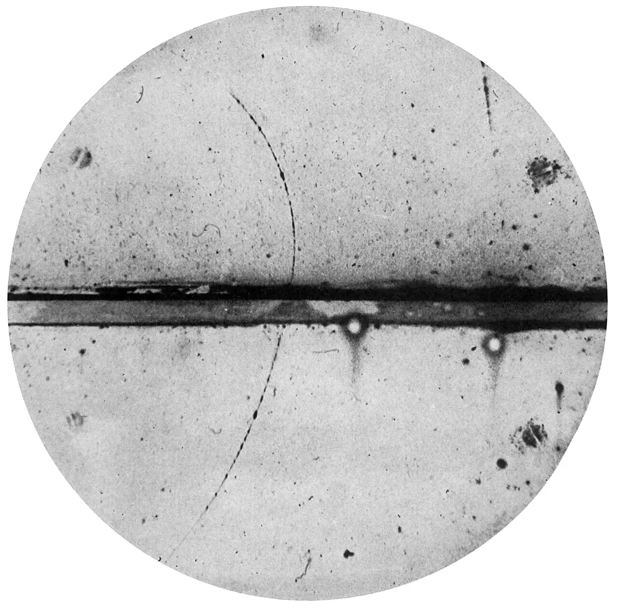 A round black and white photograph of a cloud chamber: a thin dark curving track of droplets crosses a thick dark horizontal band in the middle