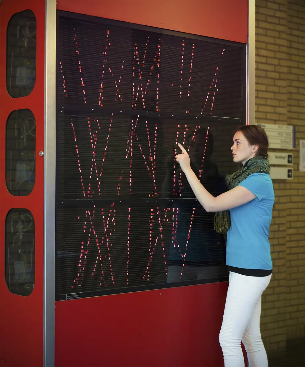 A young woman pointing at a tall glass-fronted box set in a red wall, inside which many thin red sparks form straight, slanting lines