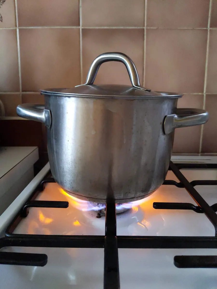 A steel cooking pot with a lid on a white gas stove; under it the flames are partly blue and partly bright yellow-orange