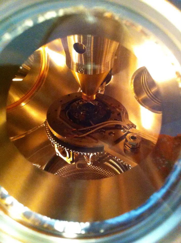 A view through a round glass window into a shiny, gold-colored metal chamber, with a pointed cone above a small sample holder