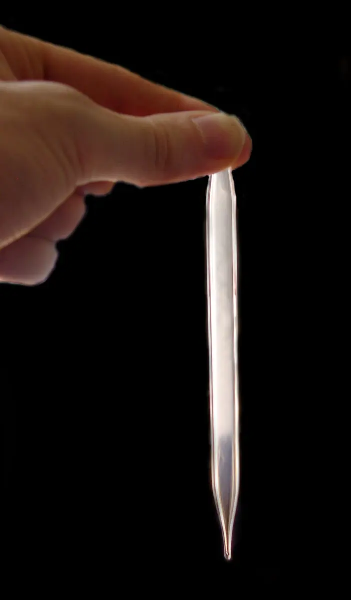Fingers hold a thin sealed glass tube, pointed at the bottom, against a black background; the fluid inside looks milky white, like a fog, and clearer near the bottom