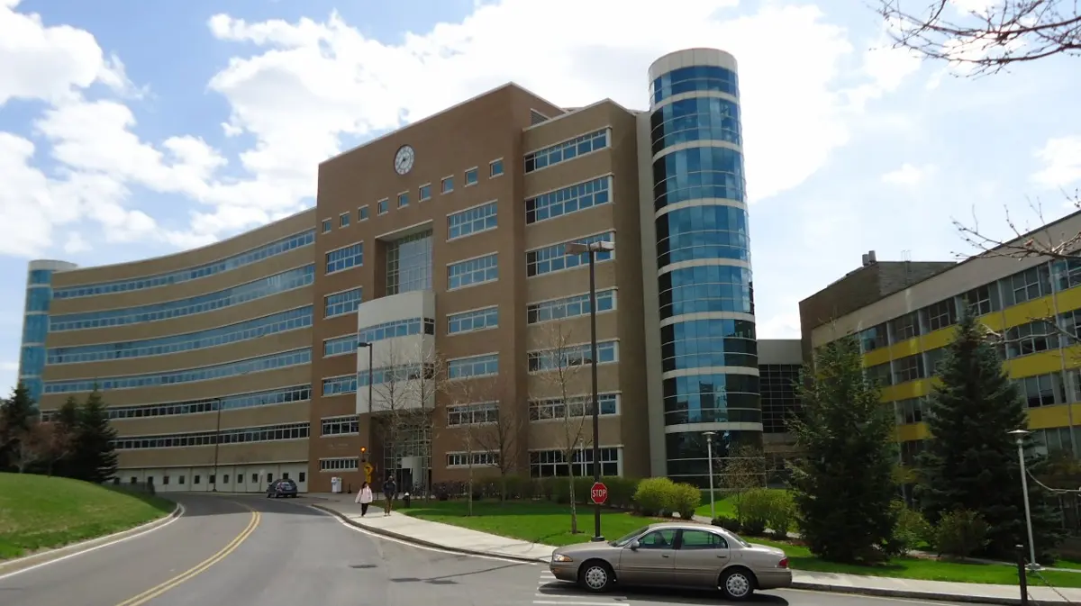 A large modern building of brown brick with long rows of windows, a rounded glass corner and a car on the road in front, under a blue sky