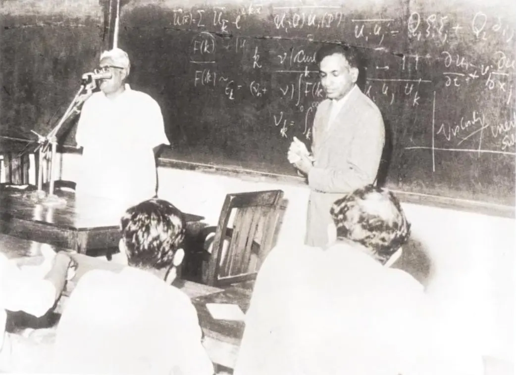 A black-and-white photo of a lecture room: a white-haired man in a white shirt speaks into a microphone on the left, while a man in a light suit stands in front of a blackboard covered in equations; people sit in the front rows