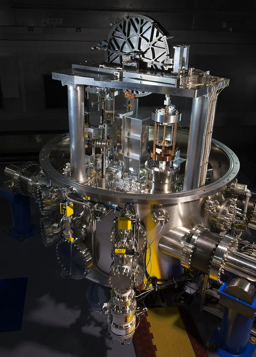 A large round steel vacuum chamber with many ports and cables, with a frame of polished pillars and a big wheel on top