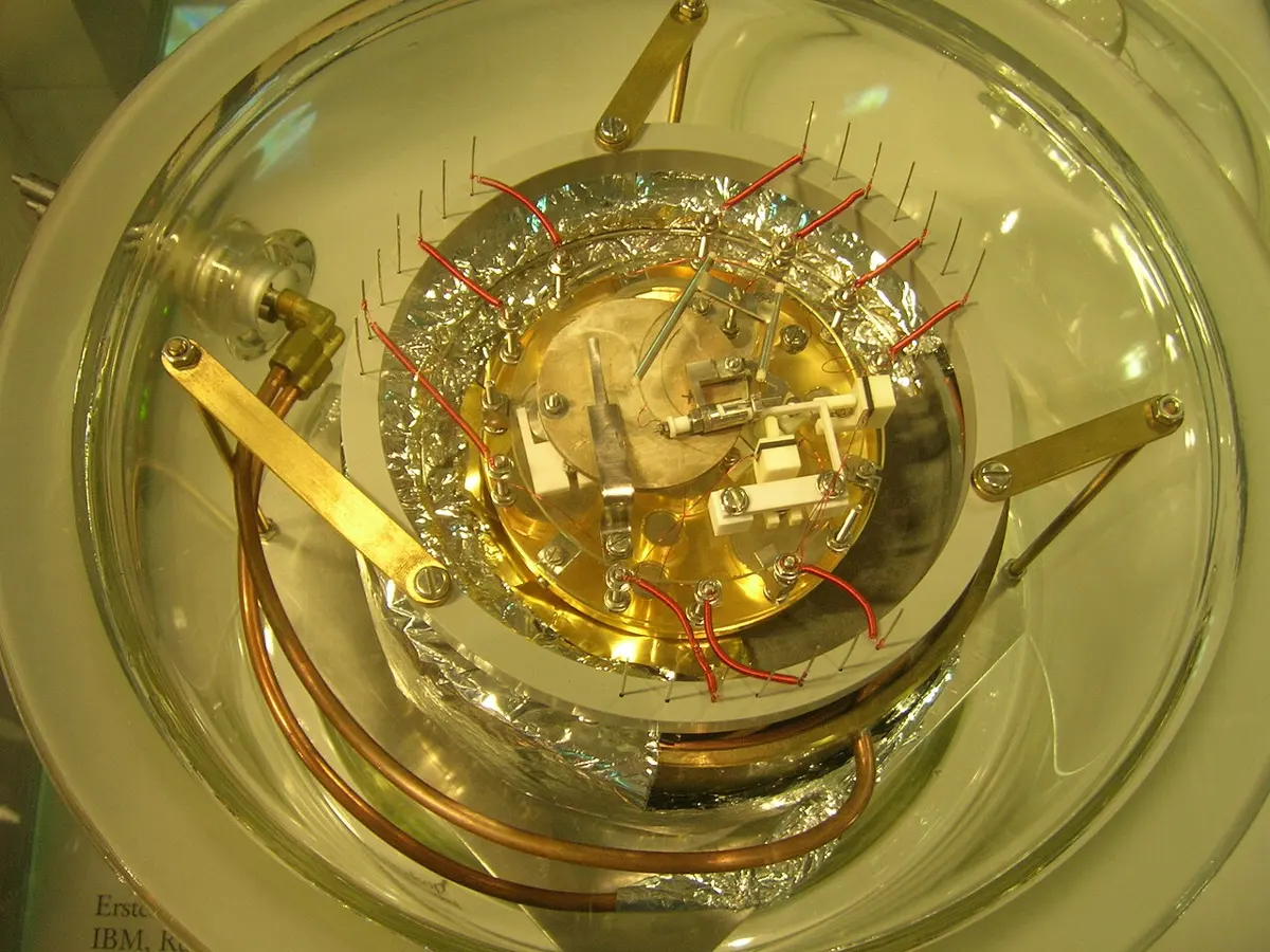 Seen from above, a round glass dish holding a golden metal plate with small parts, screws and red wires on it, sitting on crinkled foil