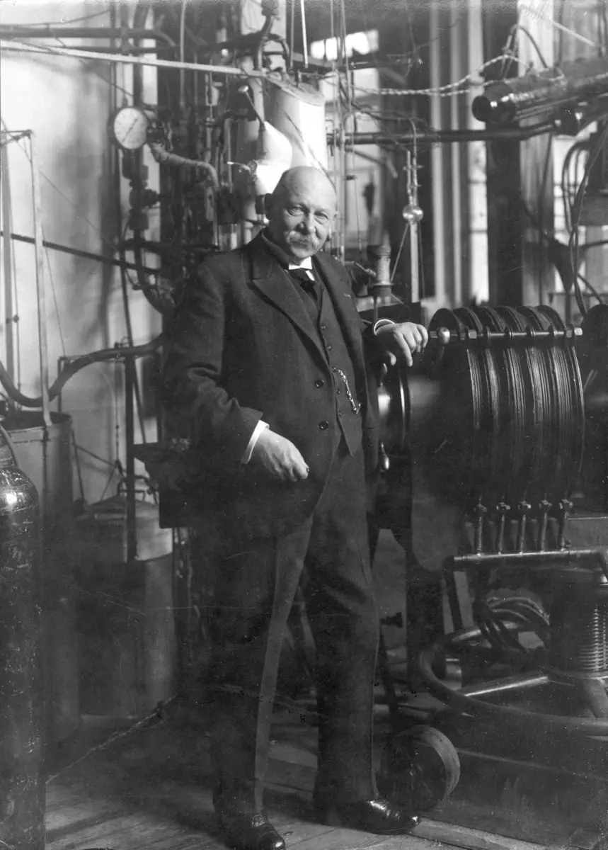 An old black-and-white photograph of an elderly man in a dark suit standing beside large pipes and machinery in a laboratory