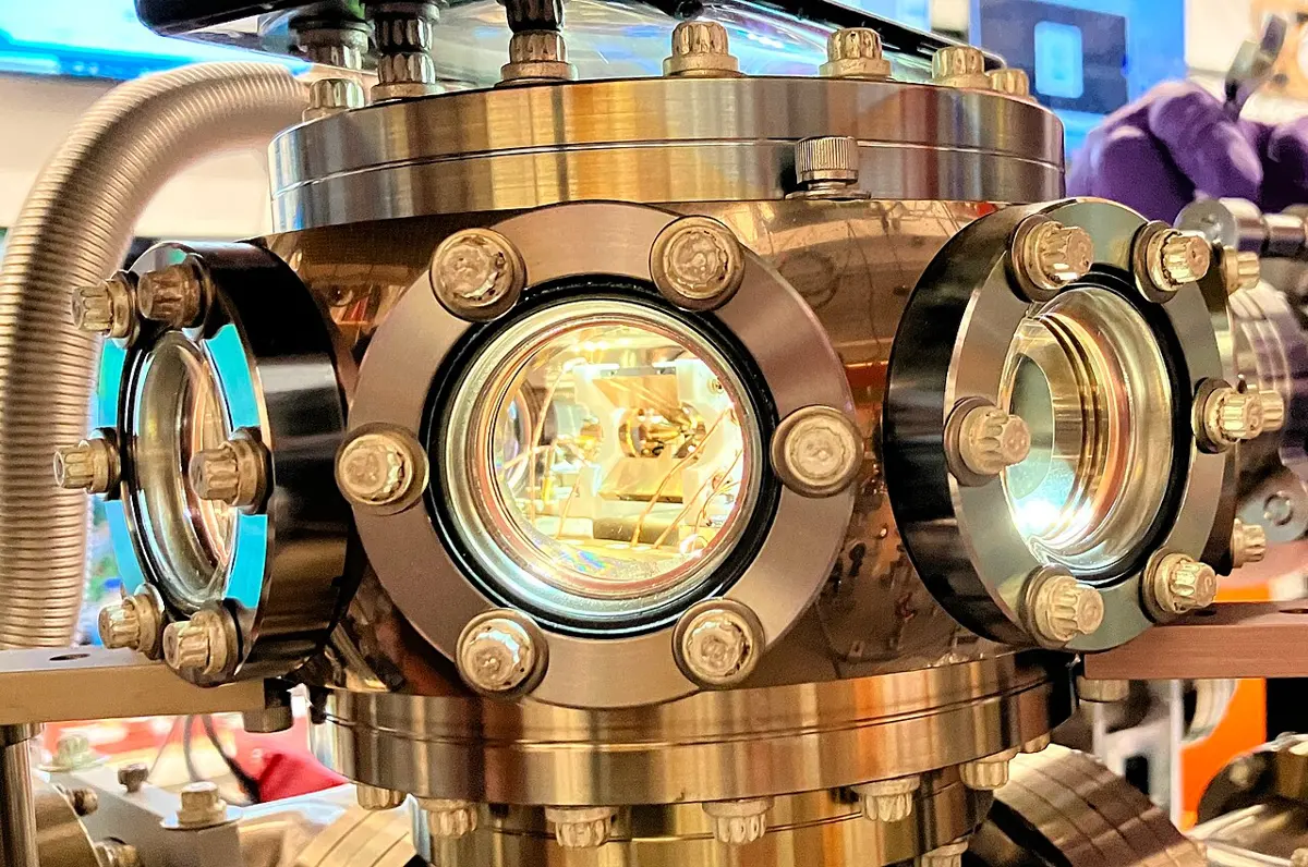 A shiny metal vacuum chamber with round glass windows held by bolts; through the middle window, small metal parts and wires glow in yellow light