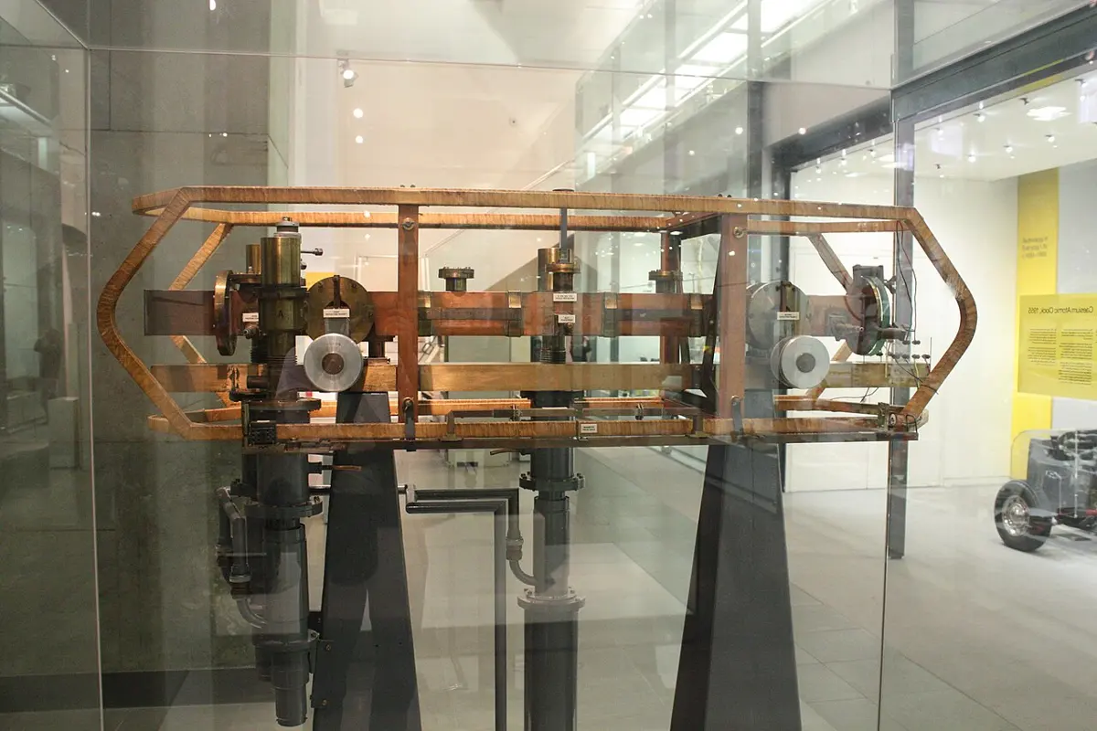 A long horizontal metal tube with brass fittings, held in a wooden frame on two stands, in a glass case in a museum