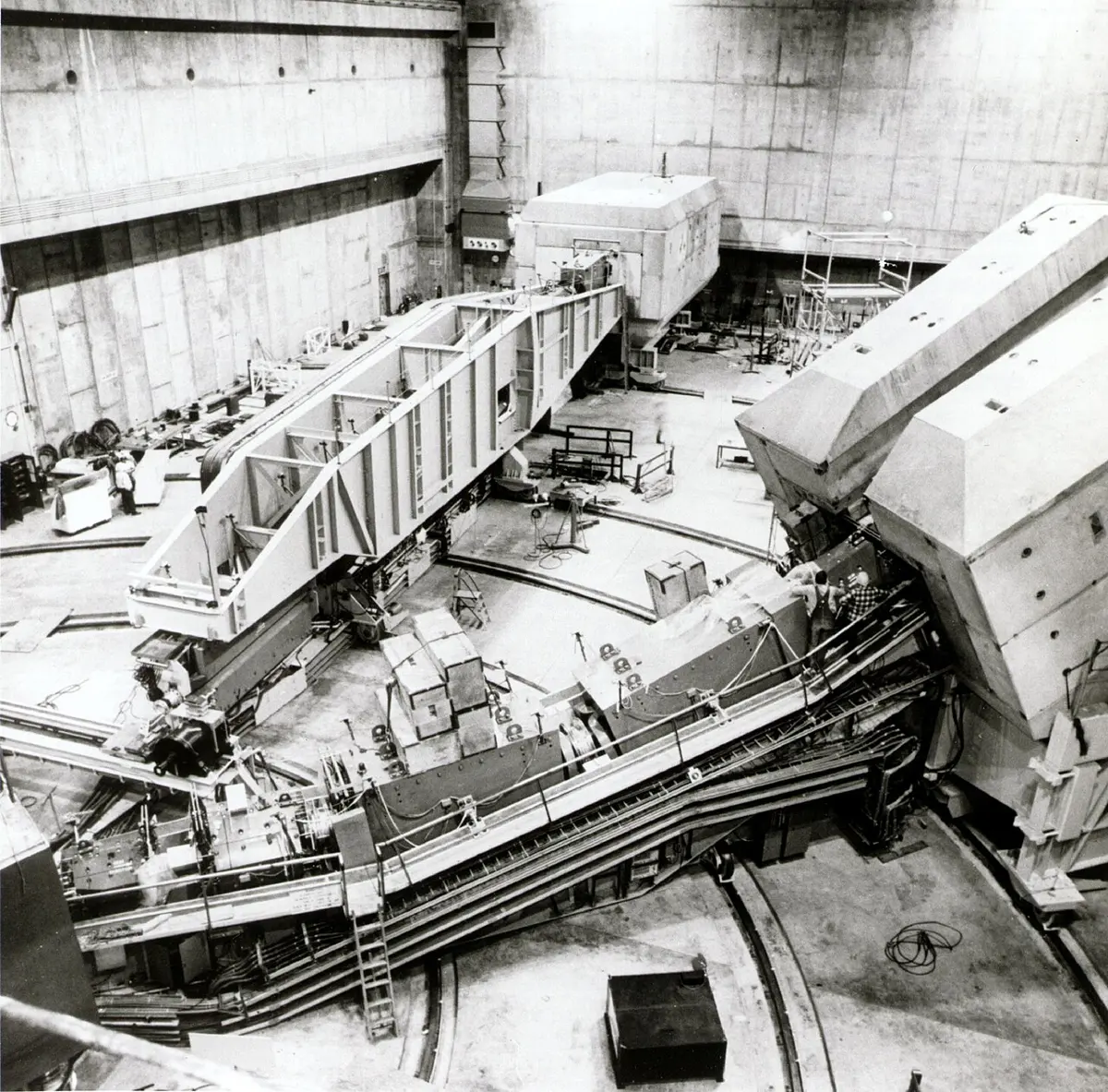 A huge concrete hall seen from above, with two enormous box-shaped machines, each as big as a house, standing on curved rails around a central point, and a tiny person standing beside one of them