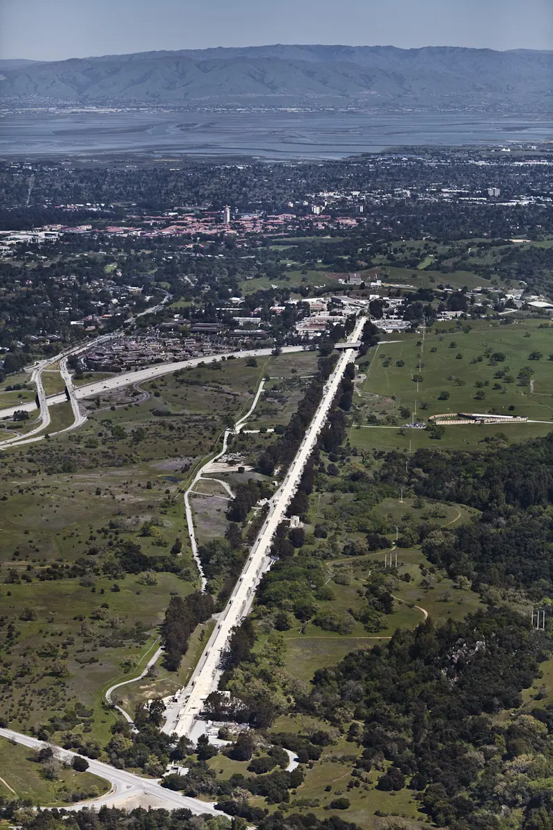 An aerial photo of a long, perfectly straight white line running for kilometers across green, hilly countryside towards a town, with a bay and mountains in the distance