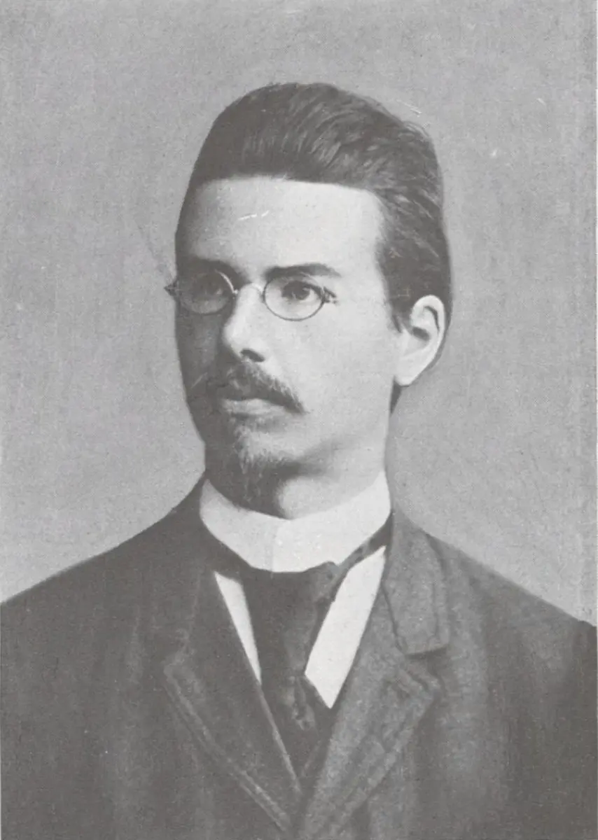 An old black-and-white portrait of a young man with swept-back hair, small round glasses, a mustache and a short beard, wearing a high white collar and a dark jacket.