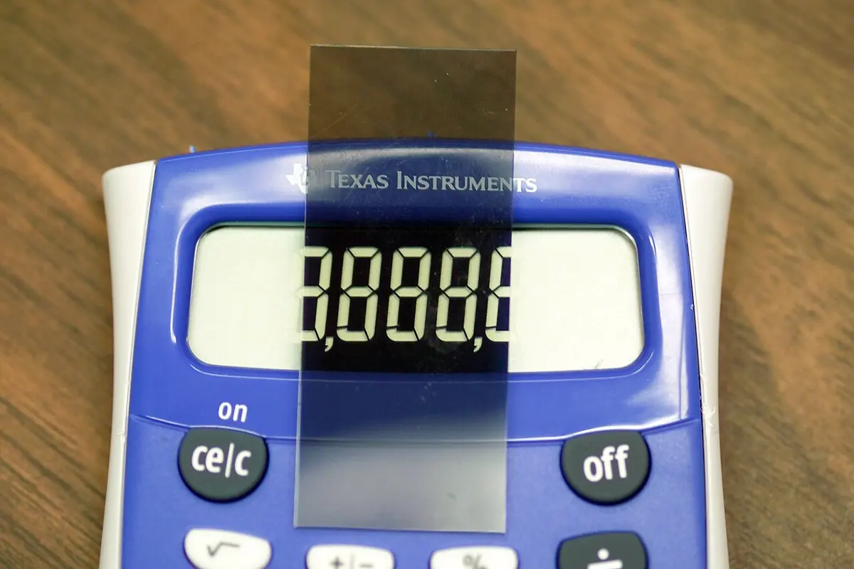 A blue calculator showing the digits 8888 in a gray display. A small dark gray strip of plastic film is held upright across the middle of the display; through it, the background looks black and the digits look light.