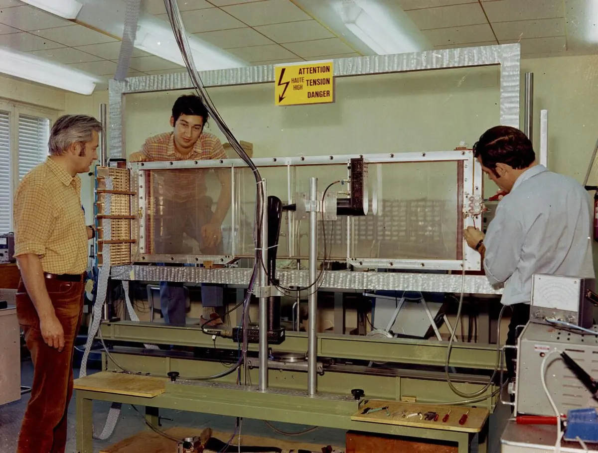 Three men in a laboratory working around a large rectangular frame with clear panels, standing on a bench, under a yellow sign warning of high voltage