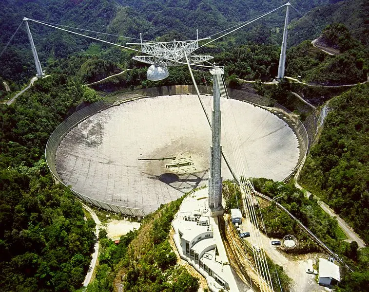 Aerial view of a huge round metal dish set into a green, hilly valley, with three tall towers holding a triangular platform on cables high above it