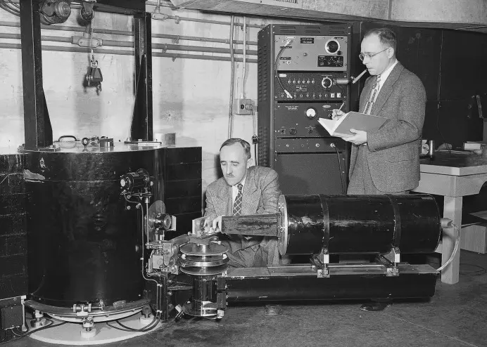 Two men in suits beside a large black cylinder and a long black tube on a stand, with a rack of electronics behind: one kneels to adjust the apparatus, the other stands writing in a notebook