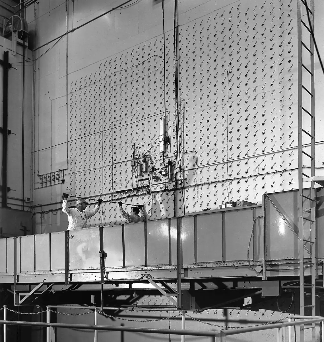 Two workers in white overalls on a raised platform in front of a huge concrete wall dotted with hundreds of small square holes, pushing long rods into the holes