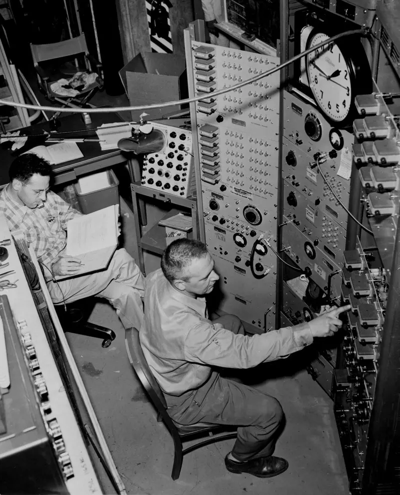 Two men in a crowded control room full of electronic panels with rows of dials, knobs and switches and a large clock: one, in glasses, sits reading a notebook; the other leans forward and presses a switch on a panel