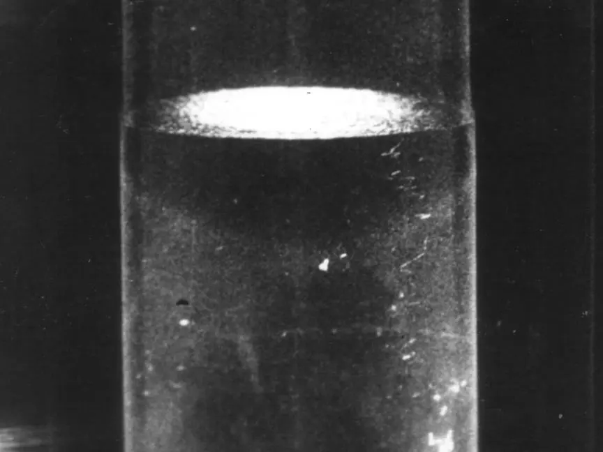 A black-and-white photograph of a tall glass vessel filled with a clear, still liquid, its smooth surface lit from the side