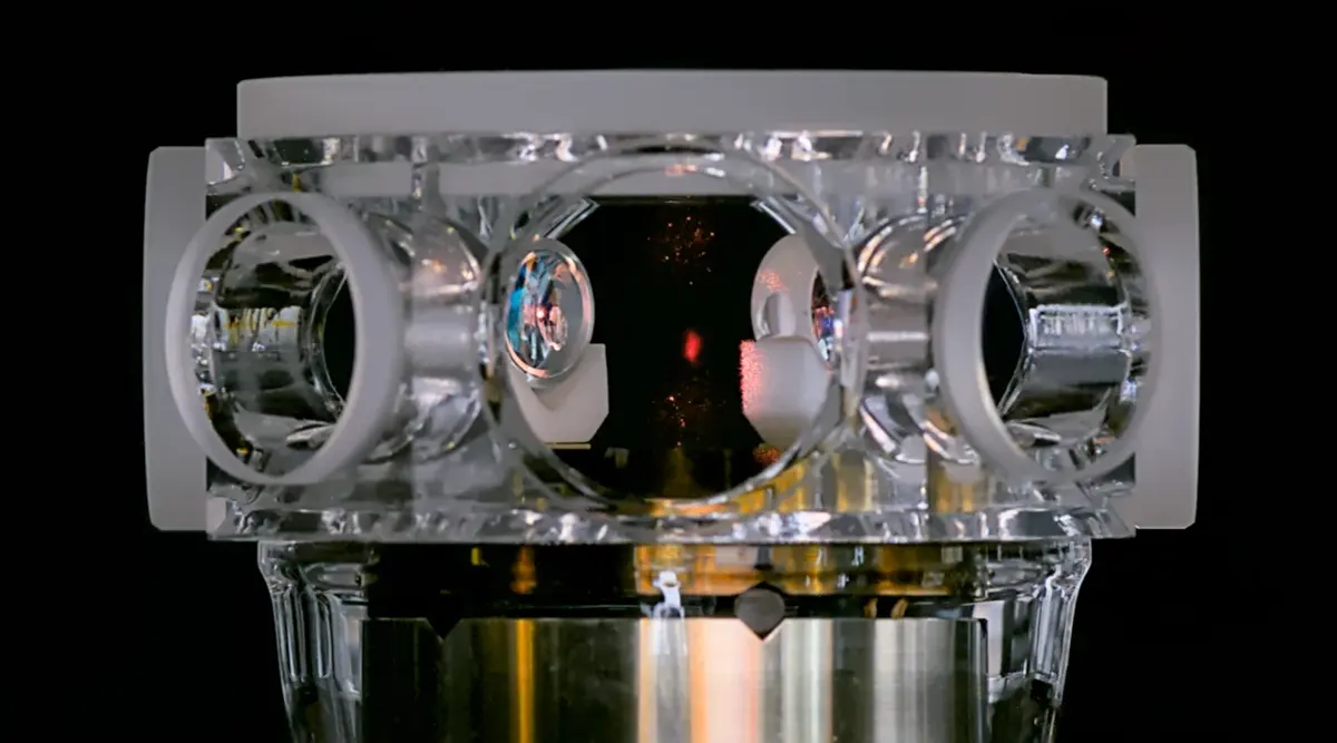 A clear glass block with round windows on each side, seen against a black background; inside, through the central window, a small red dot glows