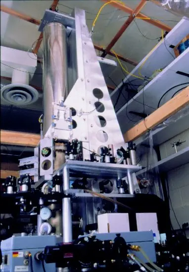 A tall laboratory instrument: a polished metal column rising from a frame on a crowded optical table, surrounded by mirrors, lenses and cables