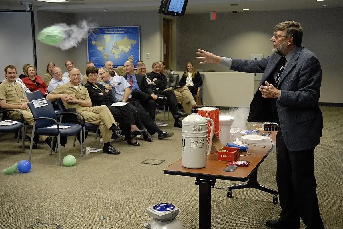 A bearded man in a suit, standing behind a table with flasks of liquid nitrogen, throws a balloon trailing white mist towards a laughing audience