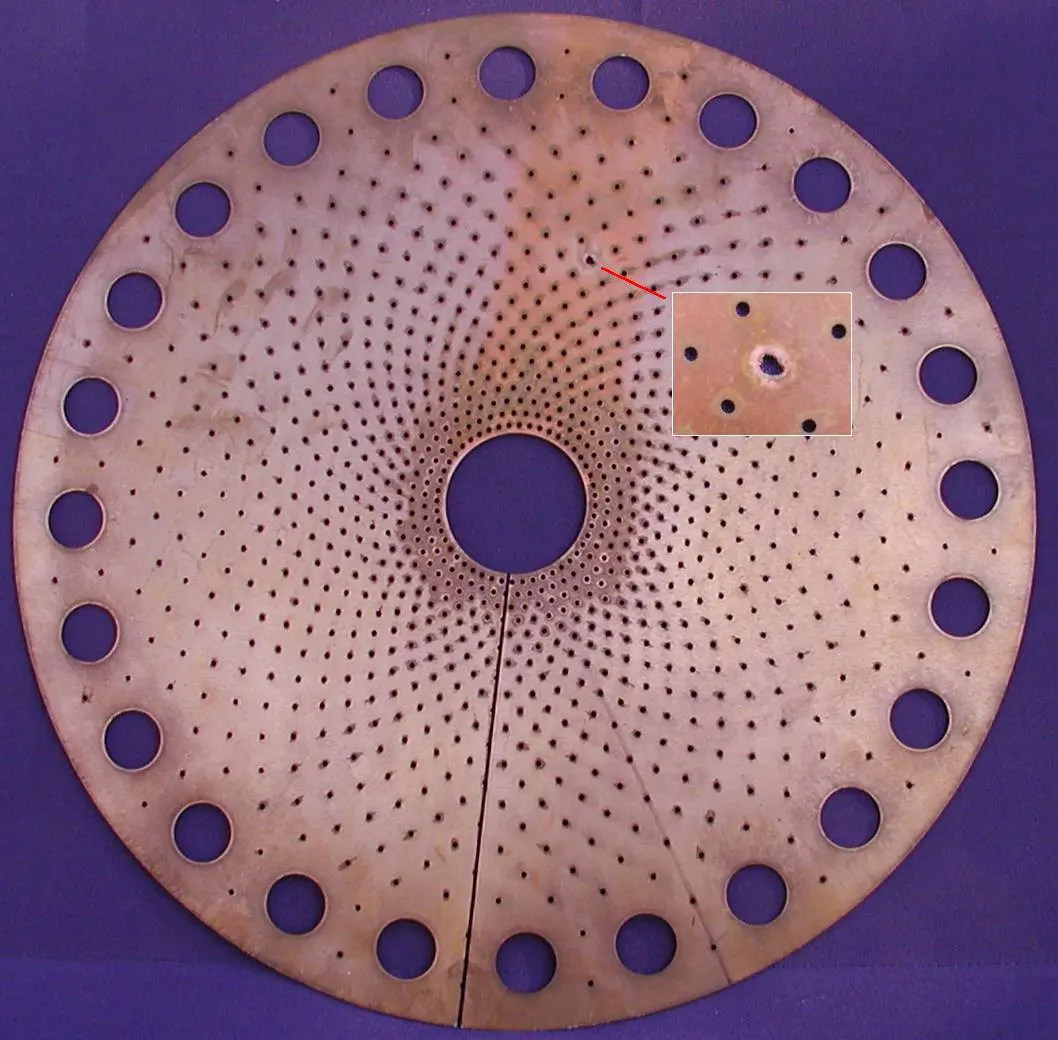 A round, flat copper-colored disc on a purple background, with a hole in the middle, a ring of large holes near its edge, hundreds of tiny holes between, and a thin cut running from the center to the edge