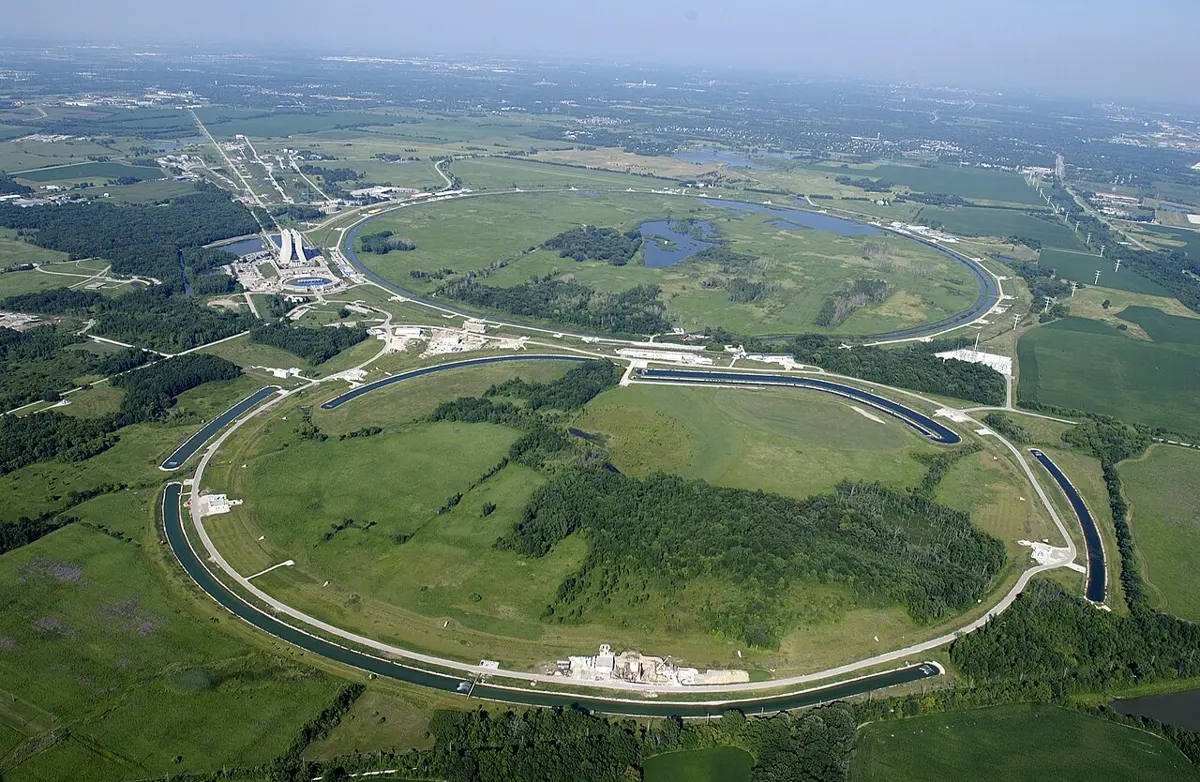 An aerial view of green countryside with two huge rings marked by roads and long curved ponds, one in front and one behind, with a tall twin-towered building beside the ring at the back