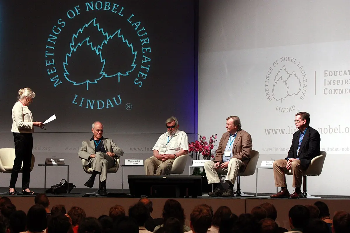 Four men seated in armchairs on a stage under a large sign reading Meetings of Nobel Laureates Lindau, with a woman standing at the left reading from a sheet of paper, and an audience in front