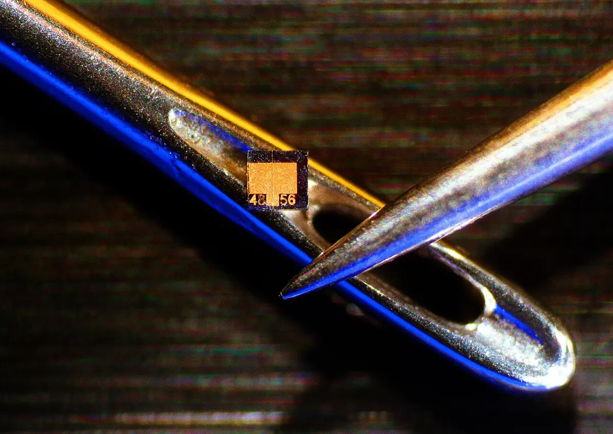 A steel sewing needle lies across the picture, and through its eye passes a second, finer needle. On the edge of the first needle’s eye sits a tiny golden-orange square chip, much smaller than the hole.