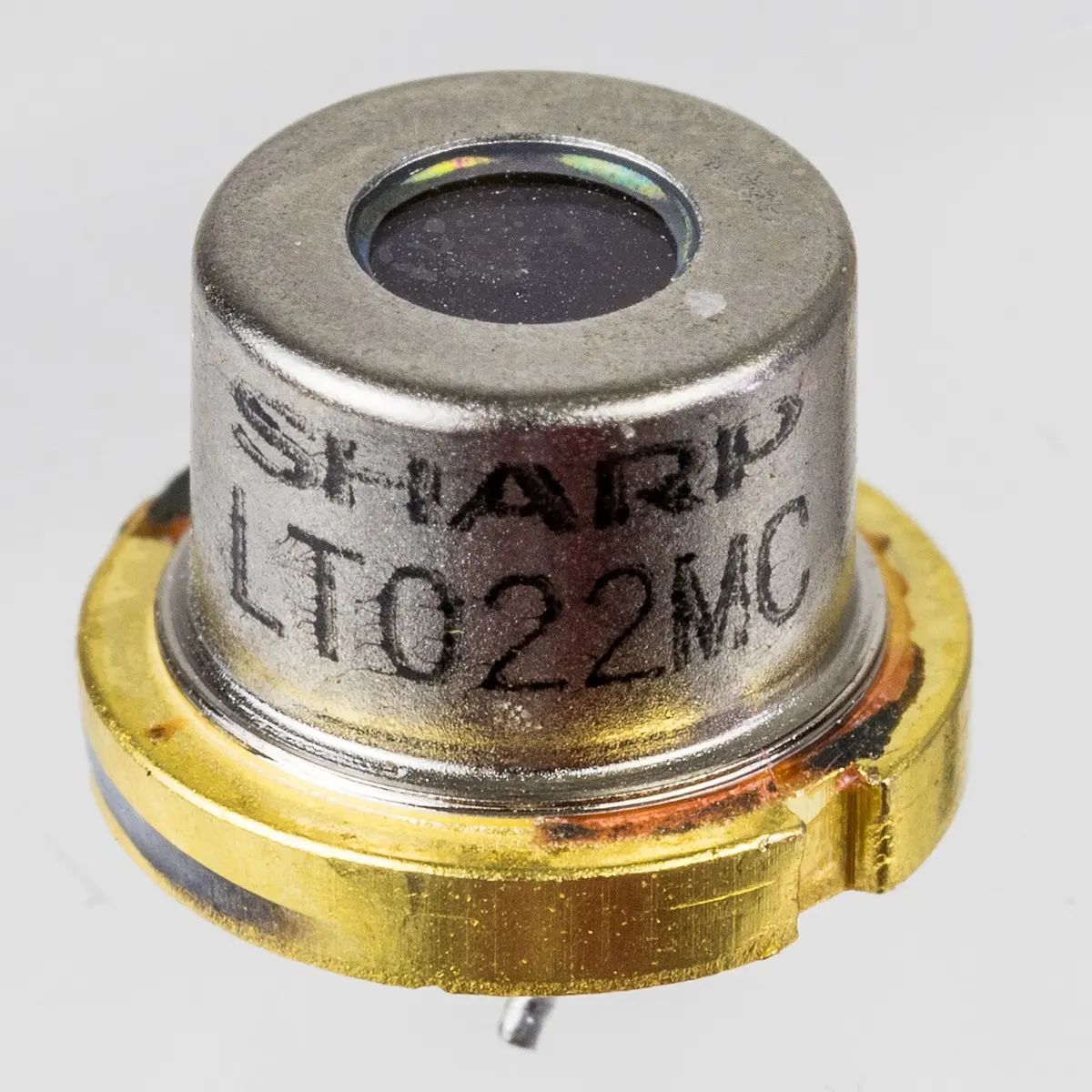 A small silver metal can, about the size of a pencil eraser, with a round glass window on top, the words SHARP LT022MC printed on its side, and a gold-colored base.