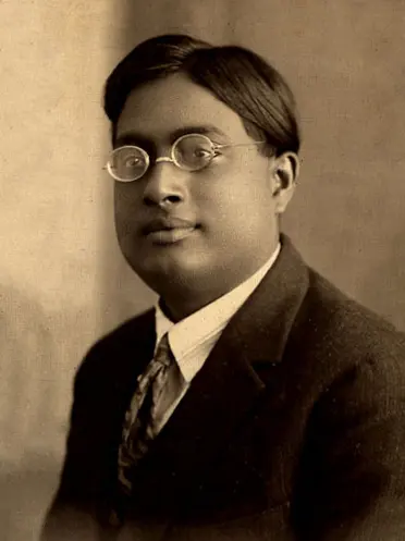 Old brown-toned photo of a young man with dark hair and small round glasses, wearing a suit and tie