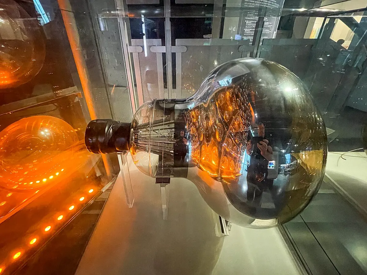 A large glass bulb, shaped like a giant light bulb about half a meter across, with a shiny mirror-like inside and a black base, in a museum glass case lit with orange light