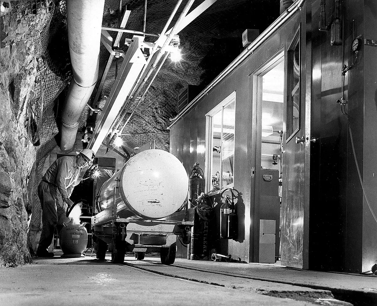 Black-and-white photo of a rock tunnel deep underground, lit by lamps, with pipes along the ceiling, a round metal gas tank on a small cart, and the metal wall and doors of a room built into the rock