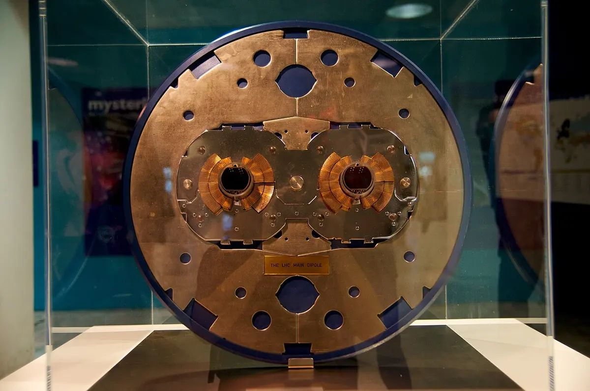 A large round metal cross-section on display in a glass case, with two holes in the middle surrounded by copper-colored coils