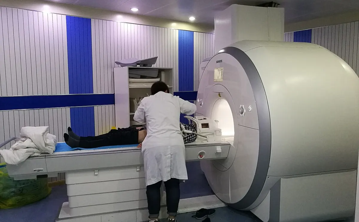 A hospital room with a large white ring-shaped MRI scanner; a nurse is getting a patient ready on the sliding bed in front of it