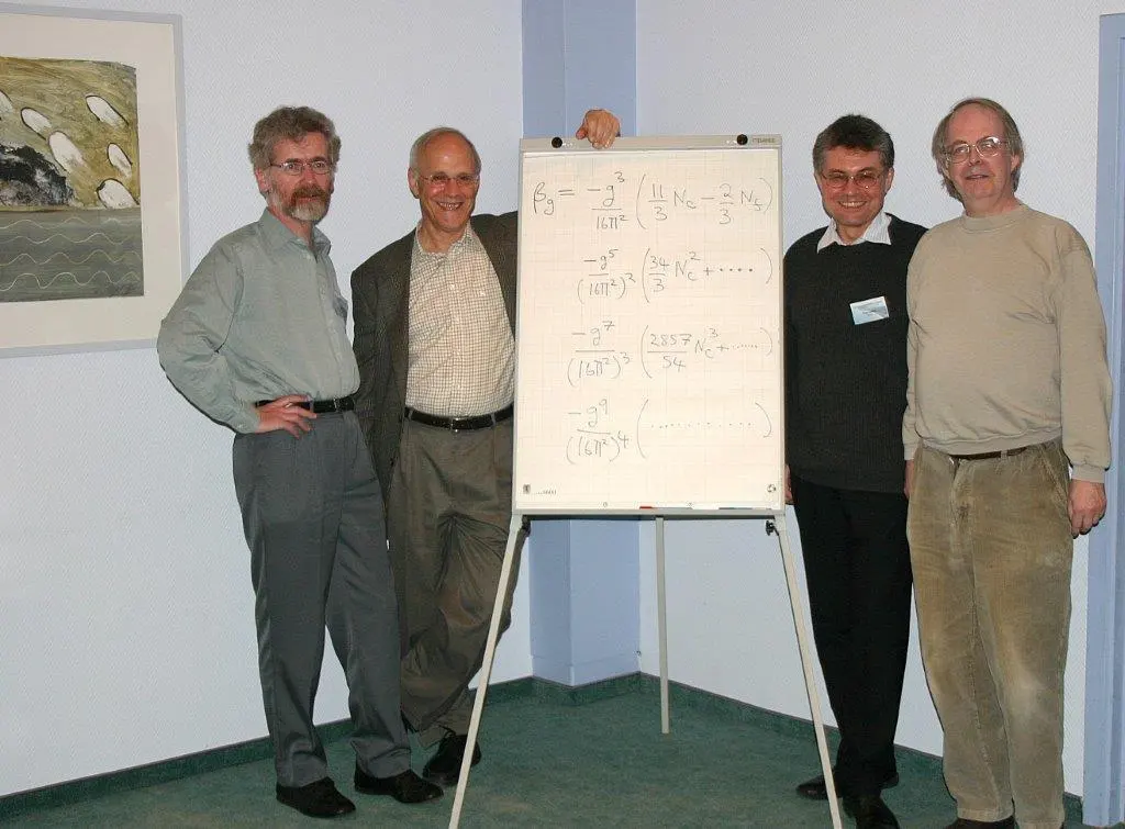Four men standing beside a flip chart covered in handwritten equations, in a room with a painting on the wall