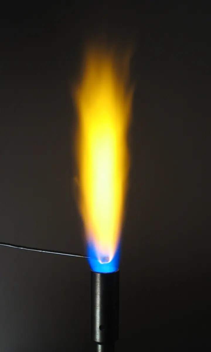 A thin wire held at the bottom of a gas burner’s small blue flame, above which a tall, bright yellow flame rises