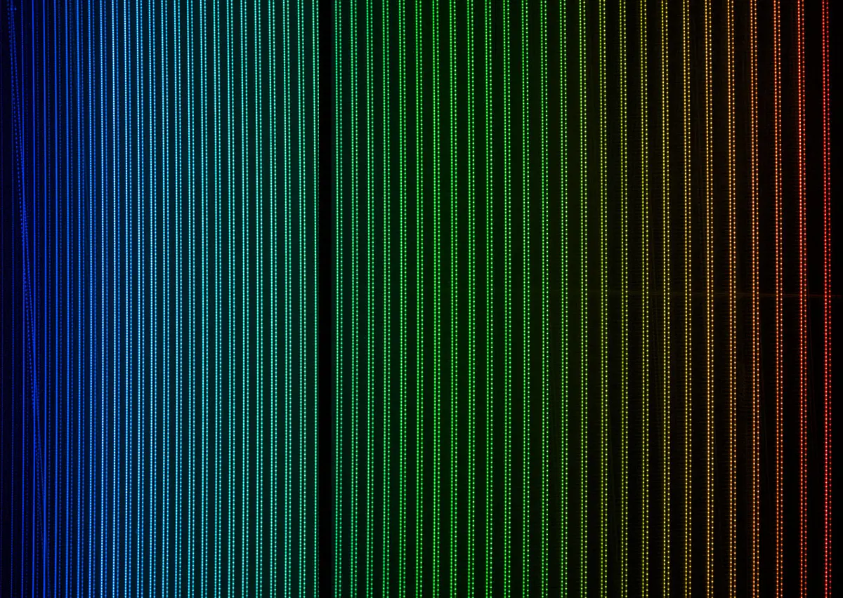 Dozens of thin, evenly spaced vertical lines of light on a black background, colored blue on the left through green and yellow to red on the right