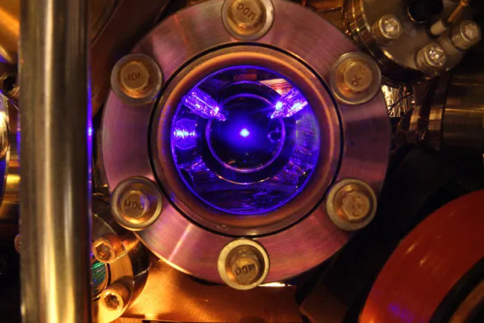 A round window in a metal vacuum chamber held by bolts; inside, laser beams cross and a tiny bright blue dot glows in the middle